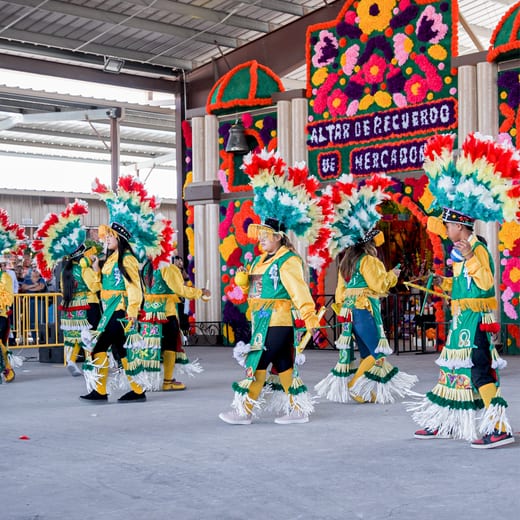 Folklórico dancers in teal costumes and feathered headdresses performing beside the market stalls.
